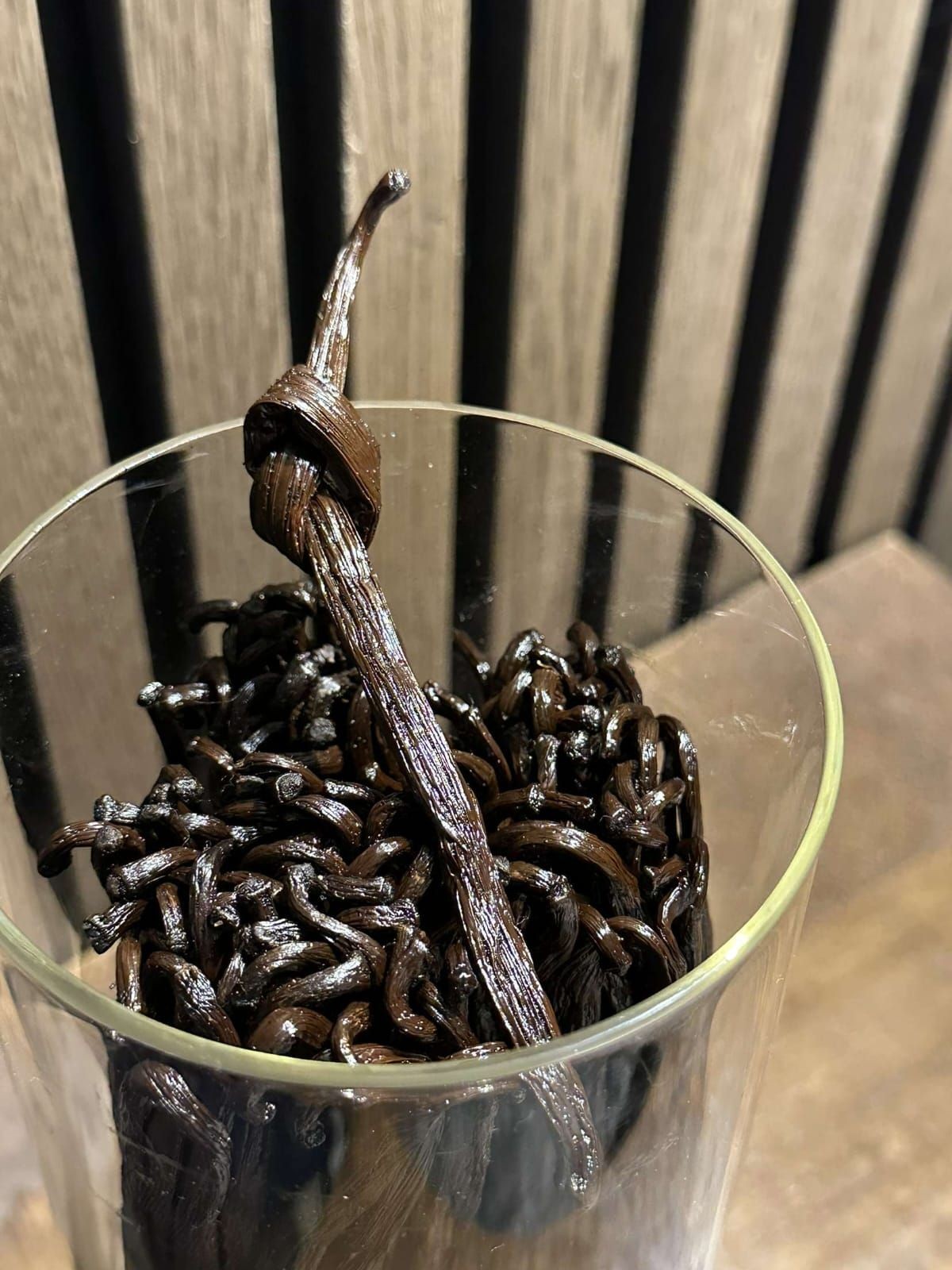 Close-up of a glass filled with shiny, dark vanilla beans against a wooden background.